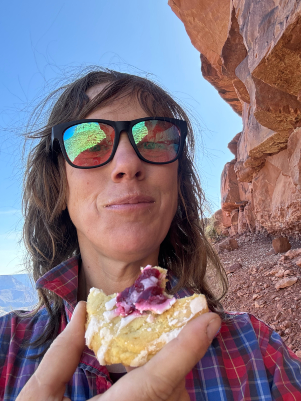 Close photo of woman in sunglasses eating a cookie in canyon country. Big rock ledge to her right, canyon walls behind her. She looks happy.